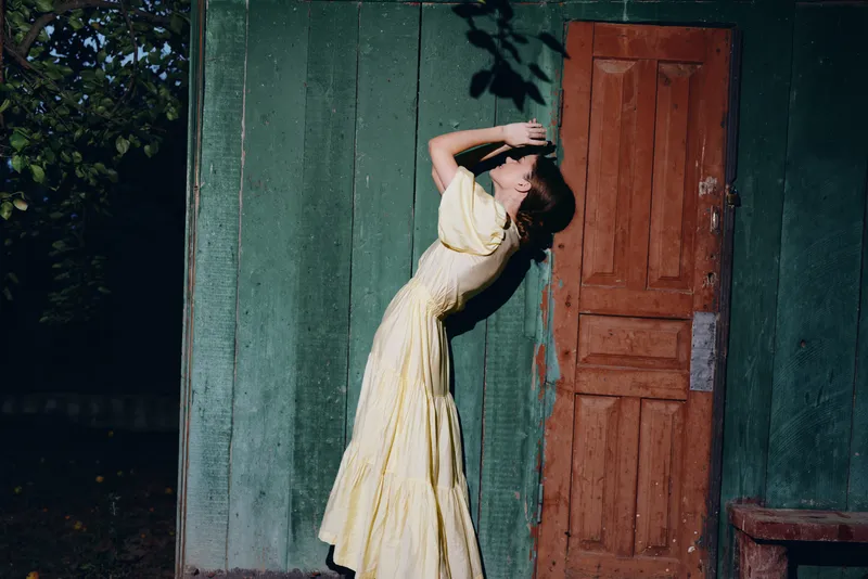 Woman in a yellow dress leans against a wooden door in an exterior yard, captured in soft light, creating a vintage outdoor moment of quiet reflection