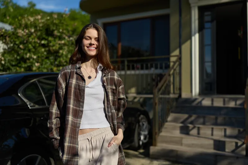 Cheerful woman outdoors in casual style, smiling with relaxed posture, sunlit street scene, natural colors, gentle breeze, greenery backdrop, candid lifestyle moment, warm and approachable