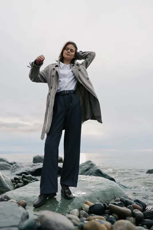 Young woman in stylish coat and trousers stands on rocky shore with cloudy sky. Casual fashion outdoors, confident pose, natural light, and sea background create moody atmosphere.