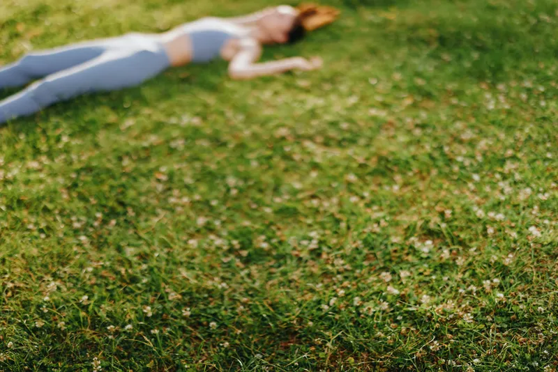 A young woman in athletic wear lies on green grass, enjoying relaxation and tranquility during a sunny day in nature, reflecting the concept of wellness and self care