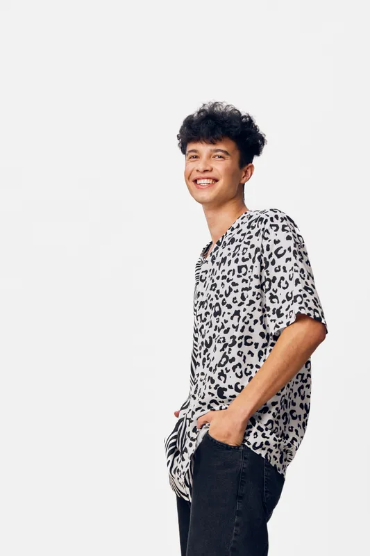 Smiling man wearing leopard print T-shirt stands relaxed against white background with hands in pockets, casual style and cheerful expression for lifestyle concept.