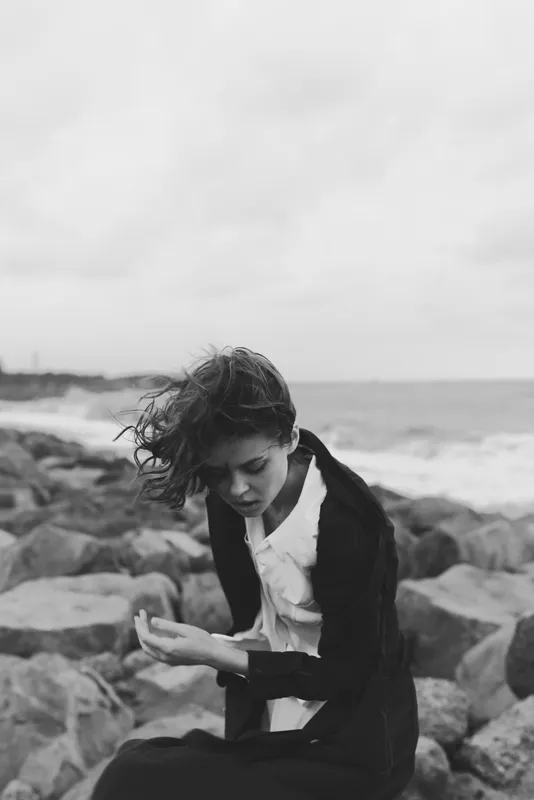 Black and white scene of a woman sitting on a rocky shoreline, looking down, wind in her hair, wearing a dark coat and white shirt, a contemplative mood by the sea with textured natural surroundings