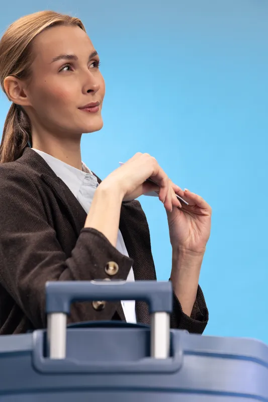 Thoughtful young woman with suitcase looks sideways on blue background. Concept of travel planning, waiting, and business trip preparation with casual outfit and natural makeup.