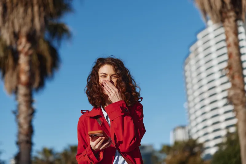 Young woman with curly hair wearing a red jacket laughs while holding a smartphone outdoors on a sunny day with palm trees and modern buildings in the background.