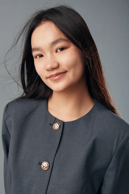 Portrait of young woman with long black hair and natural makeup in gray jacket smiling at camera on neutral background with soft lighting, confident and fresh look.