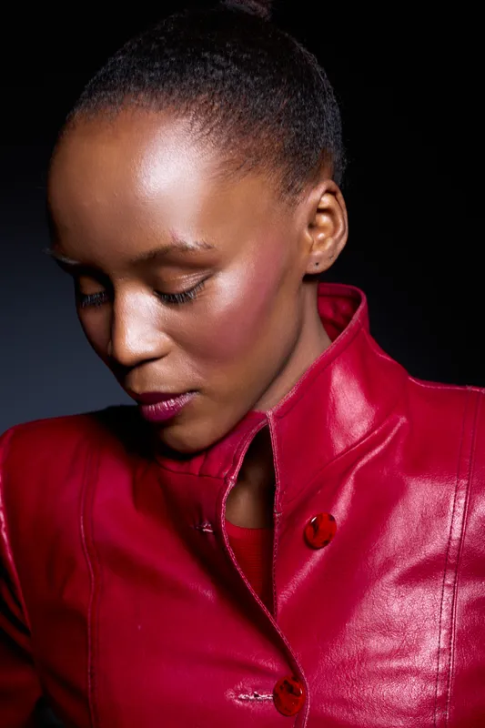 Portrait of a young African woman with dark skin wearing a red leather jacket looking down. Studio lighting highlights her smooth skin and thoughtful expression against a dark background.