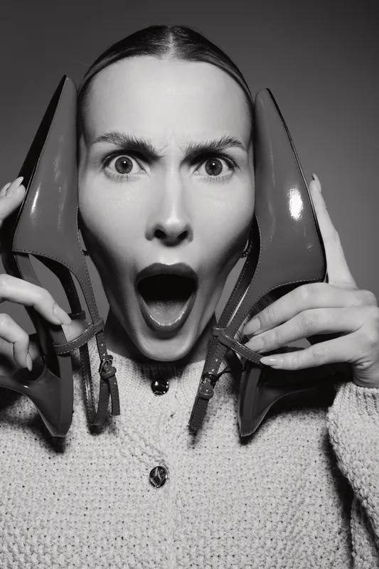 Surprised white woman with light skin holds high heel shoes near face, mouth open in shock. Studio shot with dramatic emotion, fashion and lifestyle concept.