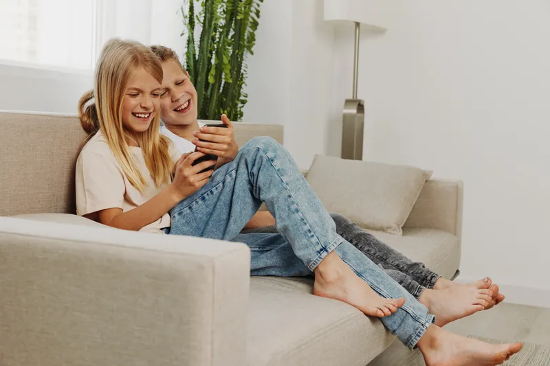 Happy children sitting barefoot on couch playing with smartphone, laughing and enjoying leisure time in bright living room. Concept of childhood, technology, family and joy.