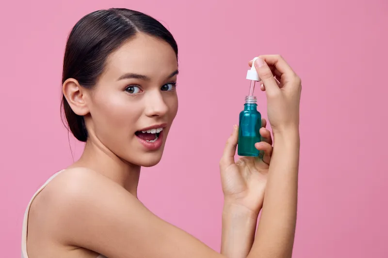 Young woman applying skincare serum, smiling and holding a dropper bottle in front of a pink background, showcasing beauty and self care routine