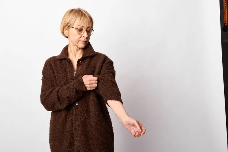 Elderly woman with glasses in brown coat shows warning gesture and refusal, serious frown, isolated portrait on white indoor background, hands half raised, tense posture