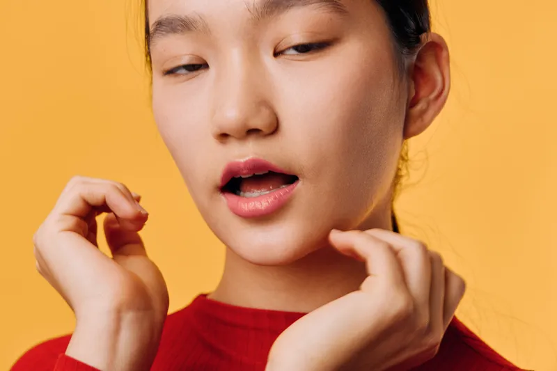 Young woman with closed eyes and open mouth posing with hands near face wearing red top on orange background. Expression, emotion and studio portrait of female model with natural skin.