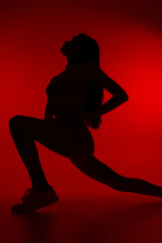 Silhouette of a woman sport fitness performing a deep lunge, showcasing strength and flexibility against a vibrant red background in activewear and sneakers.