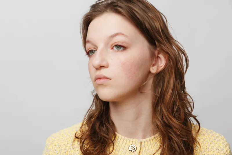 Young woman with natural skin and light freckles looks away thoughtfully against plain background. Concept of natural beauty, skin texture, and casual portrait.