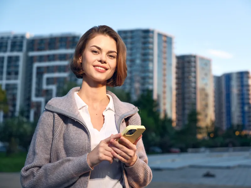 A cheerful professional woman stands outdoors in a modern urban setting, smiling as she uses a smartphone. She projects a confident, friendly, and dynamic business vibe.