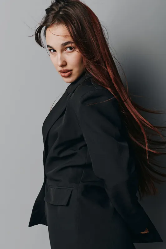 Woman with long hair wearing black blazer posing against gray wall looking confident and stylish. Female model with natural makeup and loose hair in studio portrait with soft lighting.
