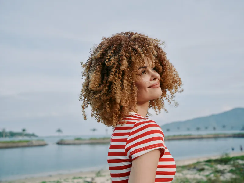 Portrait of a joyful woman with curly hair in a striped red and white shirt, standing by a calm coastal scene with a soft breeze, warm smile, and distant horizon.