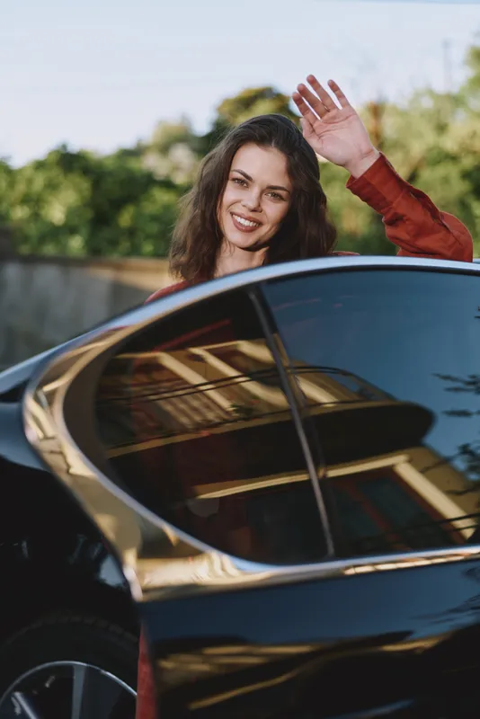 Smiling woman with curly hair waving from a car window outdoors, capturing a carefree lifestyle moment. Natural light creates a bright, energetic travel mood