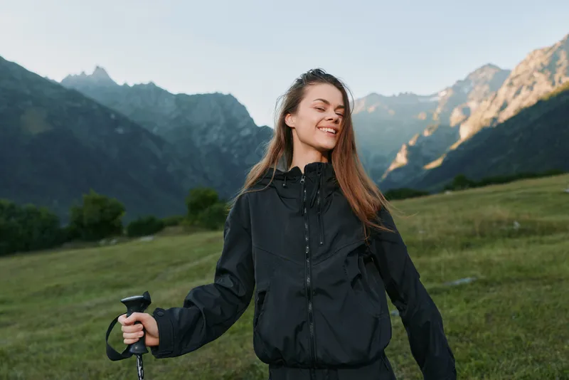 A cheerful hiker stands on a grassy hillside with a trekking pole, enjoying the fresh mountain air. She smiles confidently as rugged peaks rise in the background.