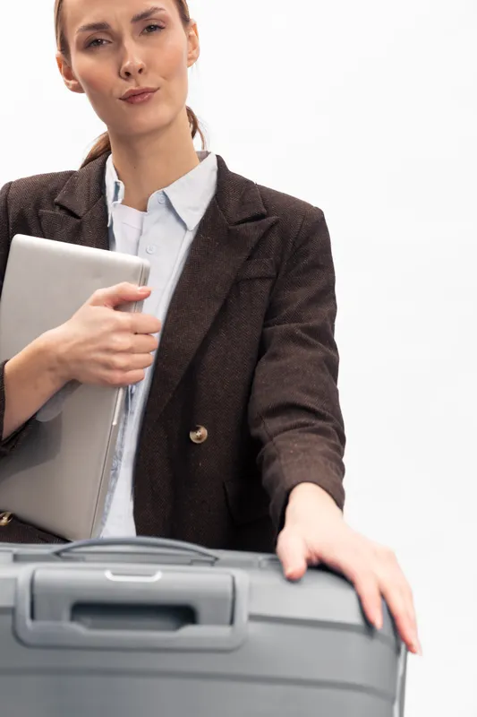 Confident woman in business attire holds laptop and touches suitcase handle, preparing for travel. Studio shot on white background. Concept of business trip, travel, and professionalism.