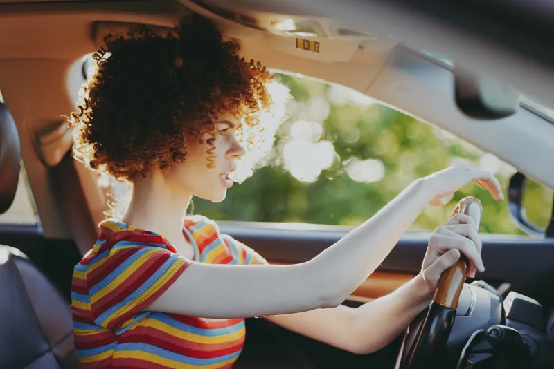 Cheerful woman driving a car, striped shirt, sunlight through the window, relaxed posture, warm retro mood, natural colors, outdoor scenery in the background