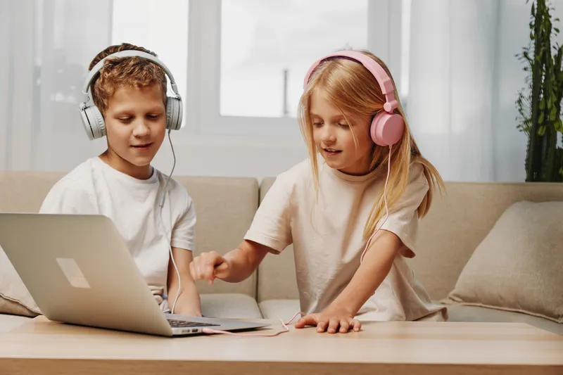 Two children wearing headphones enjoy music together while using a laptop on a wooden table in a cozy living room. Concept of childhood, technology, and shared entertainment.