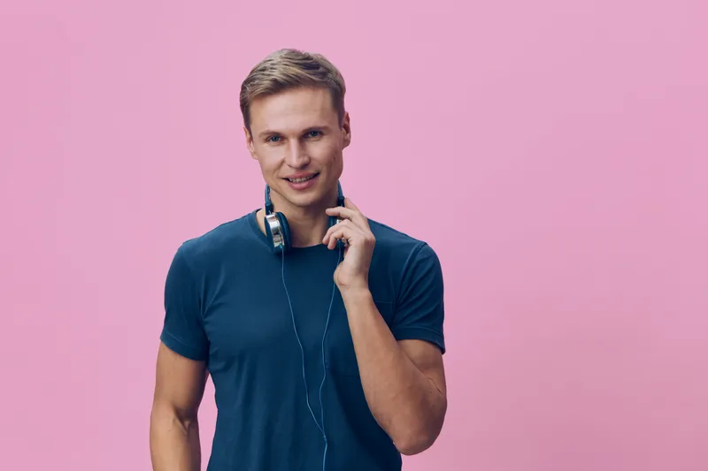 Smiling young man with short blonde hair wearing a casual blue t-shirt and headphones around his neck, looking directly at camera on pink background, portrait with happy expression and relaxed pose. P