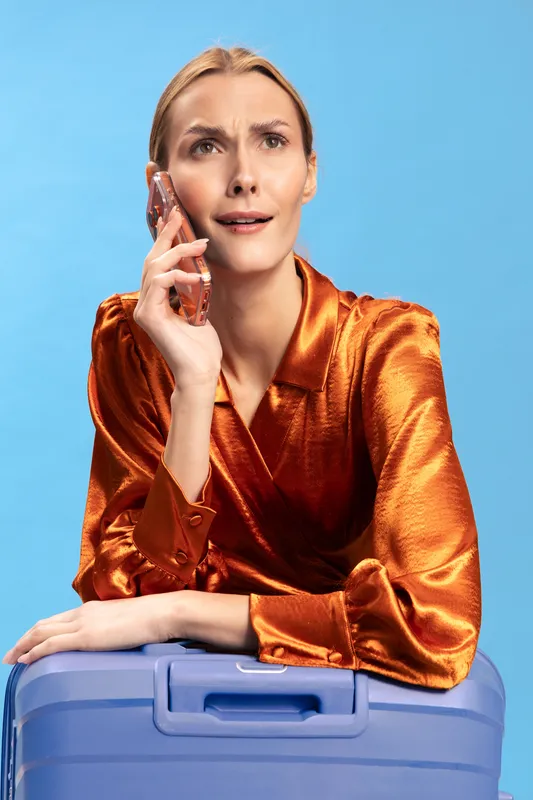 Confused woman in orange satin blouse holds phone near ear and leans on blue suitcase, expressing doubt and uncertainty. Concept for travel problems, communication, and decision making.