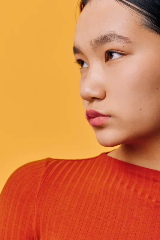 Young woman with focused face looking sideways against orange background wearing red ribbed top. Portrait of thoughtful female in warm tones with natural skin and subtle expression.
