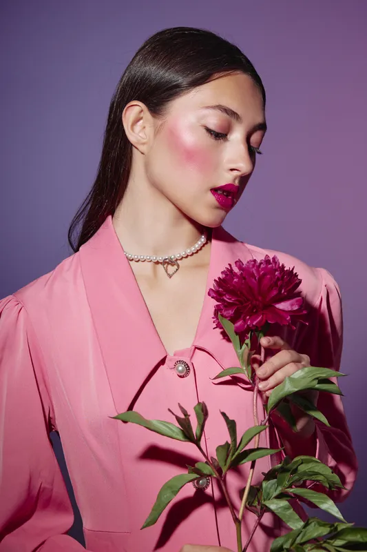 Young woman with sleek dark hair wears vibrant pink clothing and pearl necklace holding a red dahlia. Studio beauty portrait on purple background with extravagant face makeup and fashion styling.