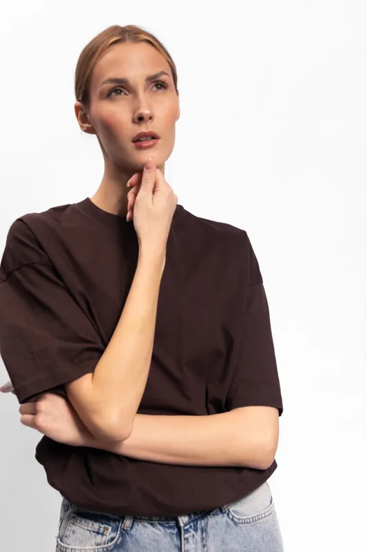 Thoughtful young woman in brown t shirt and jeans looks away while touching chin, isolated on white background. Concept of contemplation, decision making, and casual fashion portrait.