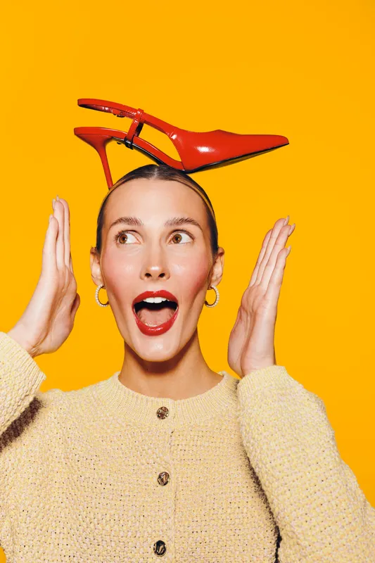 Surprised woman with red high heel shoe balanced on head looks excited with open mouth and raised hands. Bright yellow background and casual beige sweater create playful mood.