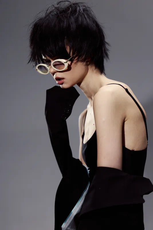 Stylish woman with short black hair wearing white sunglasses and black outfit posing thoughtfully over gray background.