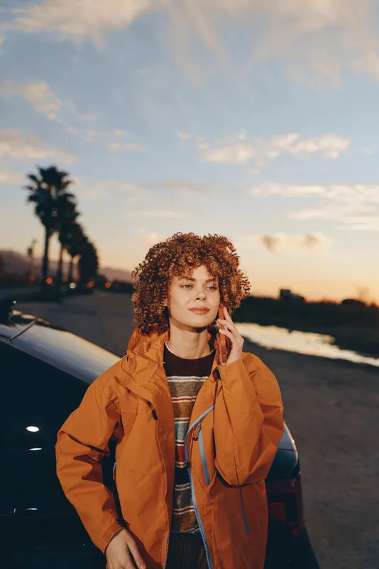 Woman lifestyle portrait outdoors by beach at sunset, curly hair under warm sky, wearing orange jacket near parked car, palm trees in background, relaxed confident mood today