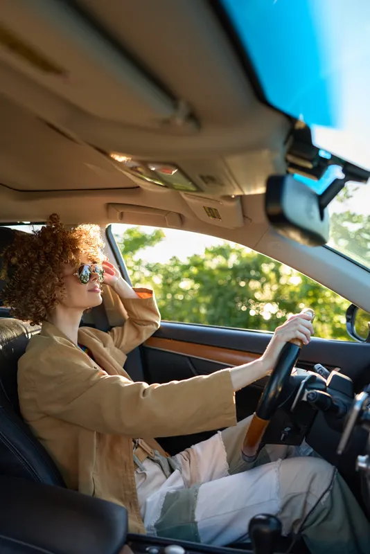 Cheerful woman driving a car, wearing sunglasses, relaxed lifestyle moment with sunlit interior and warm retro color tones, casual fashion and joyful road trip vibe