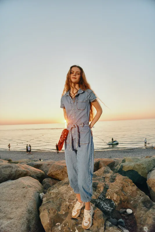 Lifestyle woman in modern boho-western denim street style stands on rocky beach at sunset with warm film color tones and relaxed confident pose