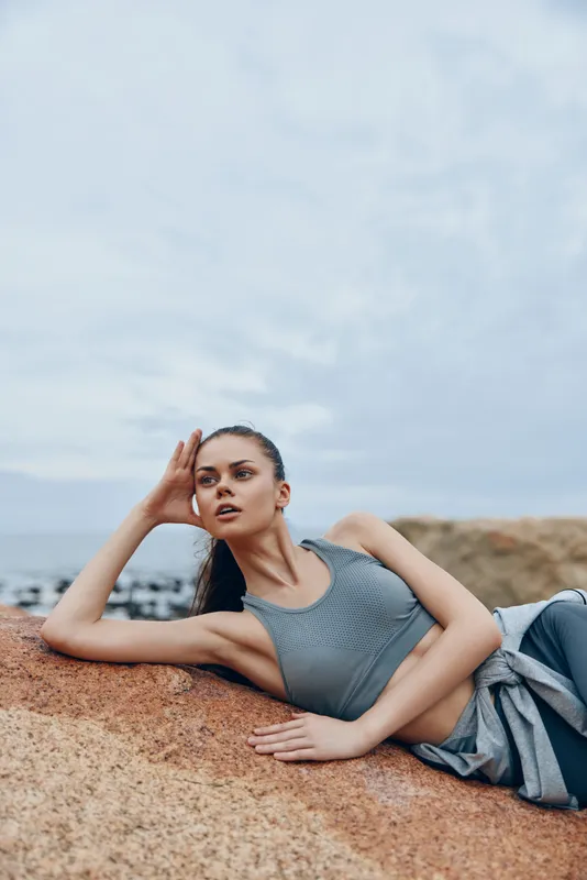 A young woman of Eastern European descent in activewear, lying on a rock with a calm expression, embodying fitness and relaxation by the seaside