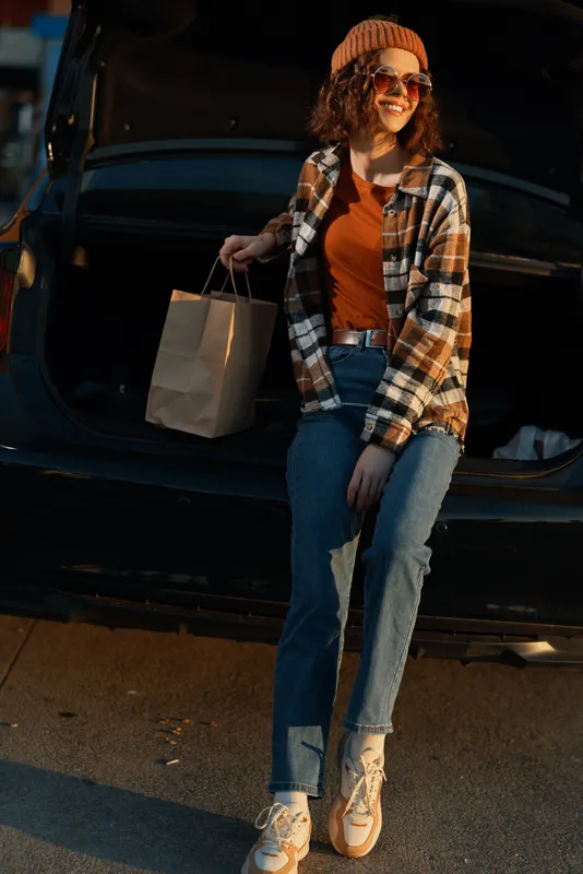 Woman car trunk shopping bag autumn casual smile sunglasses sitting on open trunk during golden hour glow, authenticity, candid lifestyle, mindful living and emotional storytelling.