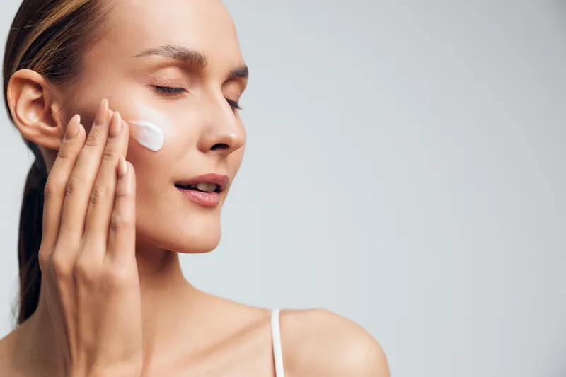 Young woman with closed eyes applies moisturizer cream on face with fingers enjoys skincare routine isolated on light background with natural fresh skin and relaxed expression.