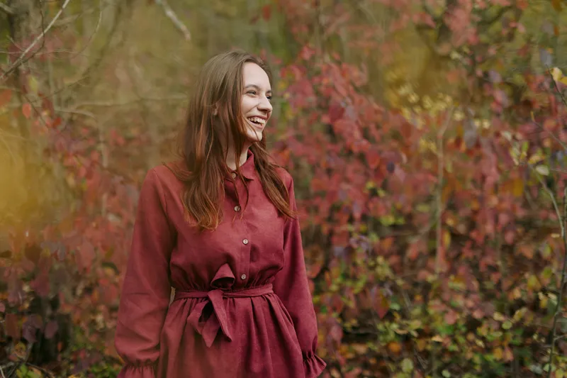 Candid portrait of a smiling woman in a rust dress amid autumn foliage, capturing an authentic expression and natural posture for a believable lifestyle scene with warm tones.