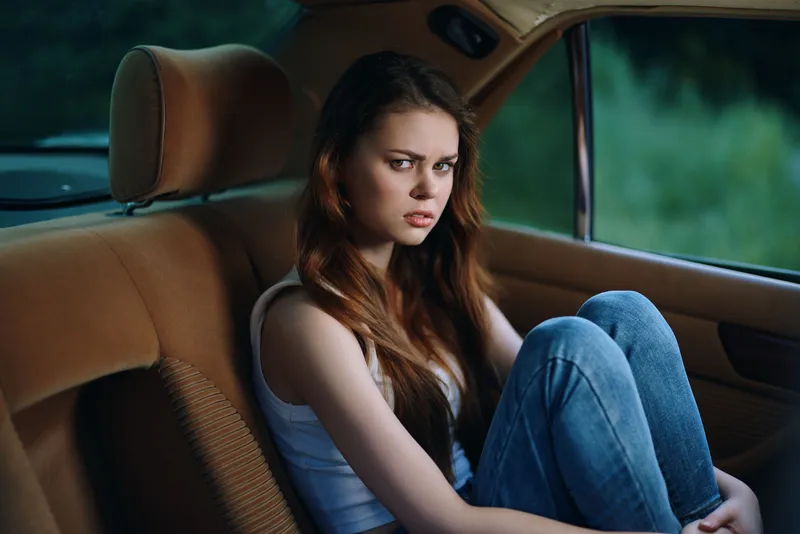 Emotional young woman with long hair sitting in a car, wearing a white tank top and jeans, expressing contemplation and introspection.
