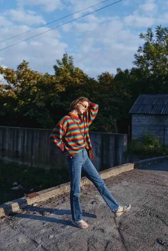 Woman, striped sweater, jeans, casual outfit, sunglasses, crossbody bag, outdoor, posing — Young woman in her mid-20s wearing a colorful knit sweater and vintage jeans, smiling and leaning on a
