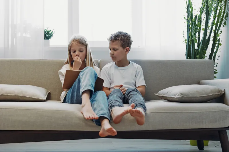 Two children sitting barefoot on a modern beige sofa in a bright living room, reading a book together with focused expressions and casual clothing.
