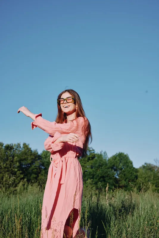 Young woman in a pink dress enjoying a sunny day in nature, expressing joy and freedom, with lush greenery in the background and a clear blue sky