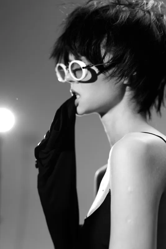 Black and white portrait of a stylish woman wearing round glasses and a sleeveless outfit with dramatic lighting.