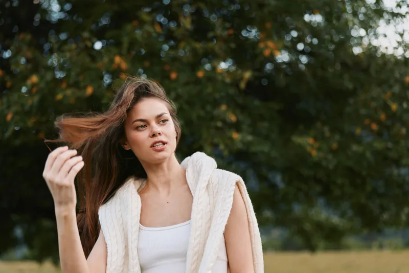 Young woman with long brown hair wearing a white tank top and knitted vest stands outdoors in a natural setting while gently touching her hair with a thoughtful expression.