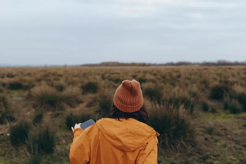 Woman outdoors in yellow coat and orange knit hat walking hiking trail across field, rear view, nature landscape and countryside lifestyle, calm fresh travel mood under cloudy sky