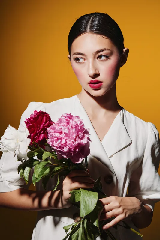 Young woman with vibrant makeup and pink lipstick holding fresh peonies against orange background. Concept for beauty, fashion, creative portrait, and floral accessories design.