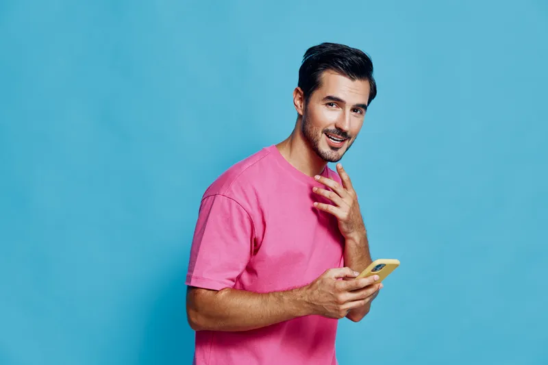 Smiling man wearing pink t-shirt looking at camera while holding mobile phone in hand against solid blue background. Lifestyle and technology concept.