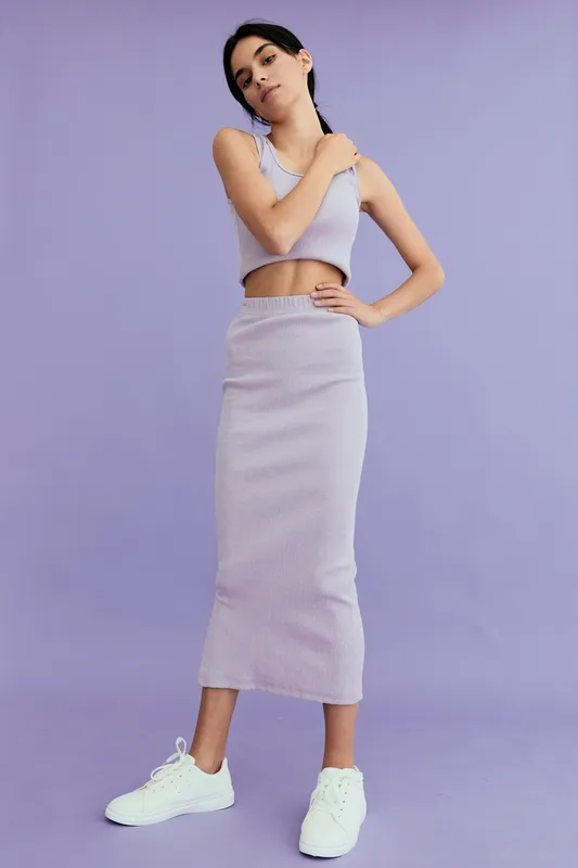 Young woman wearing a lilac ribbed crop top and skirt set stands confidently against a purple background, combining casual style with modern fashion and relaxed posture.