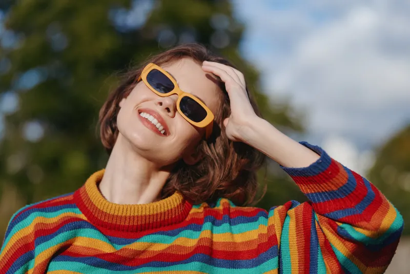 Woman smiling in a colorful striped sweater and yellow sunglasses, short hair, outdoor portrait; casual outfit, hand in hair, joyful expression, lifestyle fashion travel vibe.